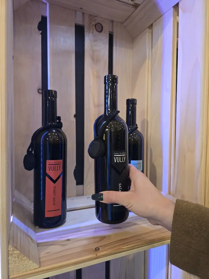 Gros plan sur la solution « lift & learn » de Hubgrade : une main soulève une bouteille de vin de la Cave Guillod munie d'un capteur de mouvement discret. Ce geste physique déclenche instantanément des informations sur les cépages et les arômes sur l'écran du stand.